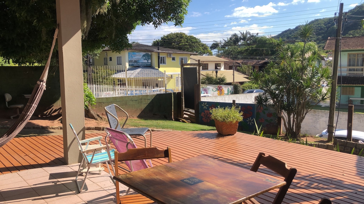 An outdoor deck area is displayed, featuring a wooden floor and various seating options, including a small table and two colorful chairs. A hammock is hung nearby, with greenery and colorful landscaping visible in the background. The view includes distant buildings and a blue sky.