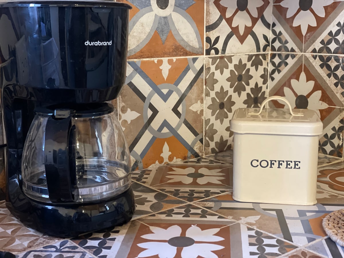 A black coffee maker is positioned next to a cream-colored coffee canister on a tiled countertop. The tiles feature intricate patterns in warm tones, contrasting with the modern appliance, creating a functional kitchen space.