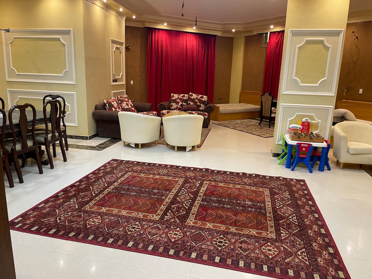 A spacious living area features a large, patterned rug that anchors the room. Comfortable seating is arranged around the space, with a combination of armchairs and sofas. Rich, red curtains frame the area, enhancing the warm tones of the decor.