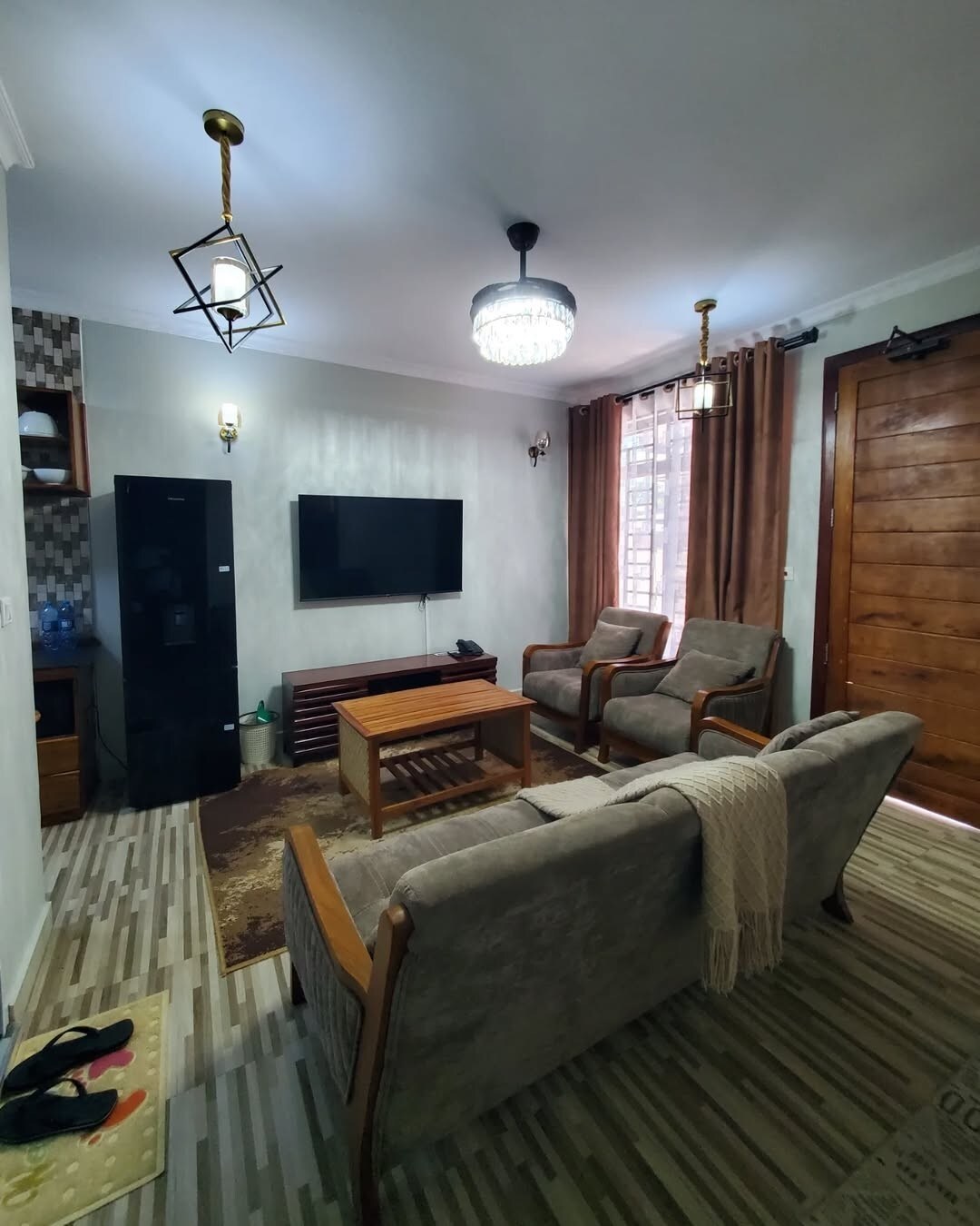 The living area features two comfortable armchairs and a sofa arranged around a wooden coffee table. A flat-screen television is mounted on the wall, and a rich wooden door leads to the entrance. Natural light filters through sheer curtains, illuminating the space.