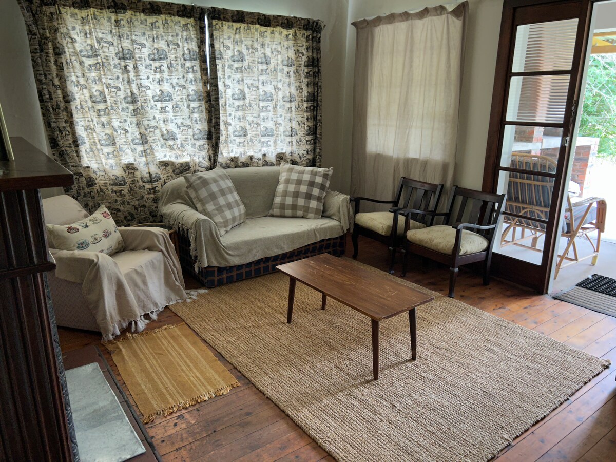 The living area features a cozy sofa with checkered cushions and two armchairs arranged around a wooden coffee table. Natural light filters through patterned curtains, illuminating the space. A woven rug adds texture to the wooden floor, creating a welcoming ambiance.