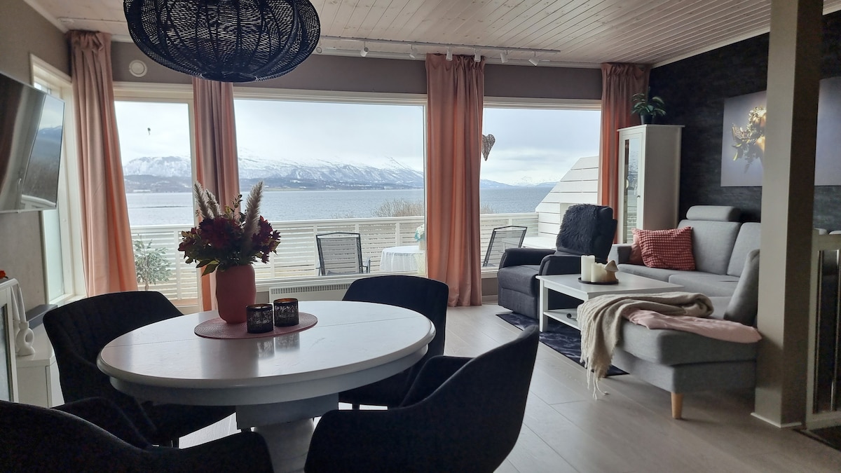 A dining area is presented with a round table surrounded by black chairs. Large windows offer views of the landscape, while light filters through flowing curtains. A cozy seating area is styled with a grey sofa and decorative accents, creating an inviting space.