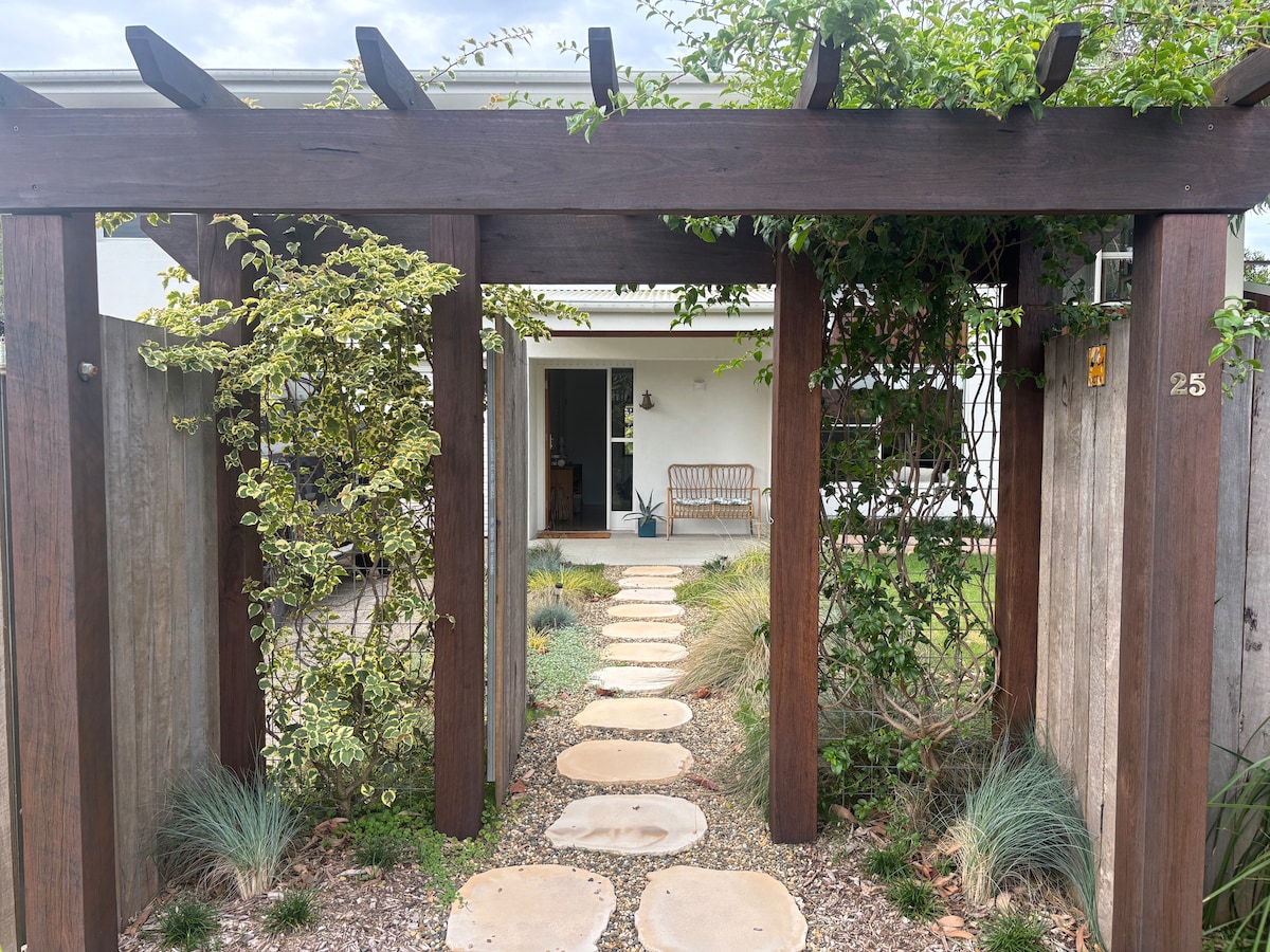 A welcoming entrance is framed by wooden beams, leading to a serene path of smooth stones. Lush greenery borders the walkway, with a glimpse of a light-colored house in the background, inviting guests to explore the tranquil surroundings.
