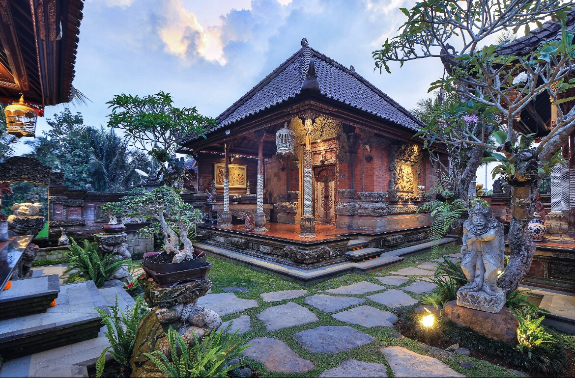A traditional Balinese structure features intricate stone carvings and a pointed roof, surrounded by lush greenery. Stone pathways lead to the entrance, with potted plants and statues enhancing the serene ambiance. Soft garden lighting illuminates the area, creating a tranquil outdoor space.