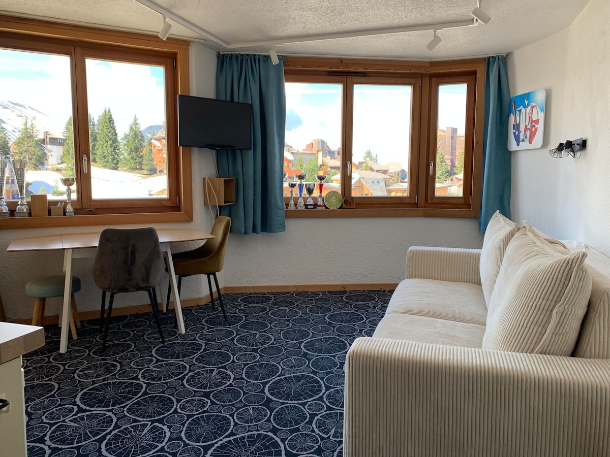 A bright living area features large windows with mountain views, allowing natural light to fill the space. A cozy sofa is positioned next to a small dining table and chairs, with a TV mounted on the wall nearby. The floor is covered with a patterned carpet.