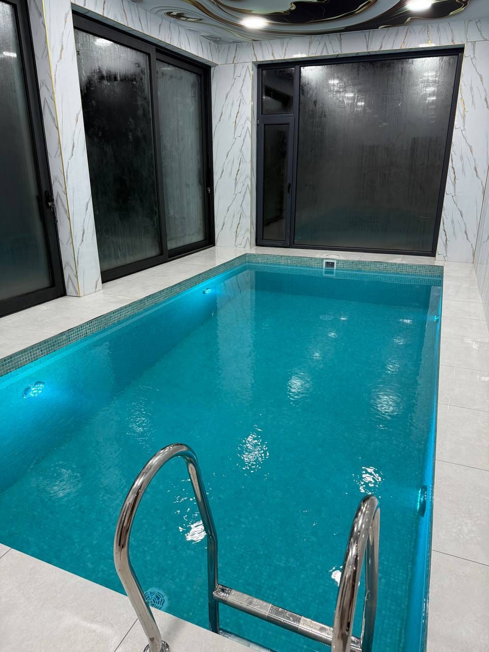 An indoor pool is featured, surrounded by sleek marble walls and large glass windows allowing for natural light. The pool's water is a clear turquoise, with steps that lead down into the inviting space.