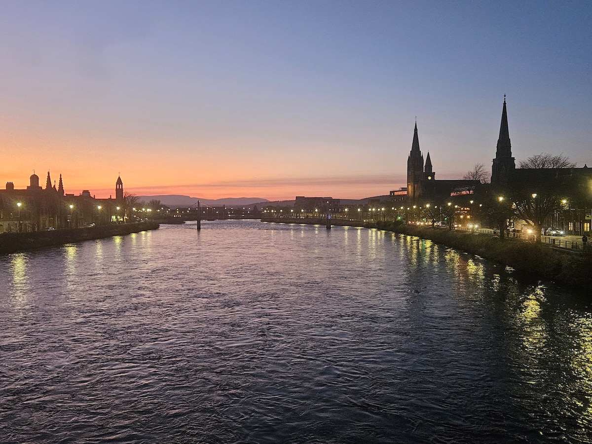 A serene riverside view captures a twilight scene with a vibrant sunset reflecting on the water. Silhouetted buildings line the waterfront, including spires and clock towers, while the warm glow of streetlights begins to illuminate the surroundings.