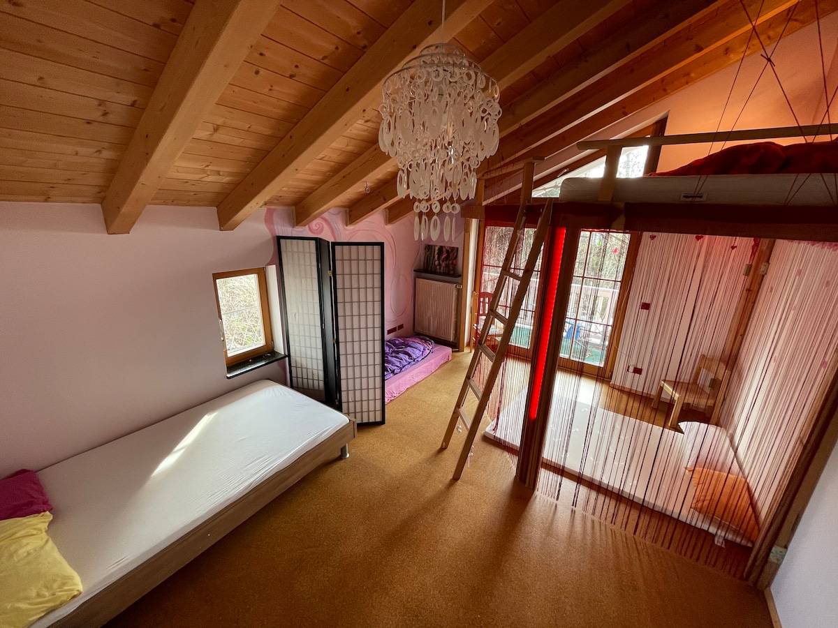 An overhead view of a room with wooden beams and a high ceiling. A single mattress is positioned on one side, while two comfortable air mattresses in soft colors occupy another area. A ladder leads to a loft space, and a decorative screen divides sections of the room.