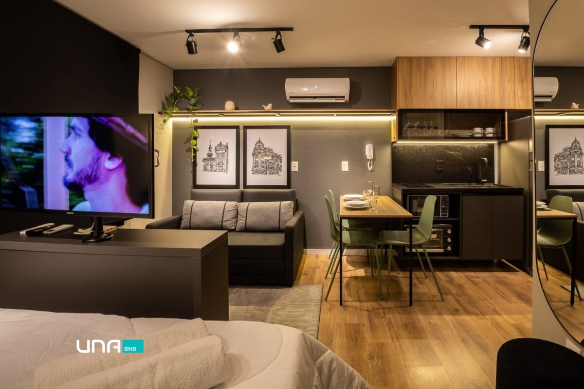 A modern living area is presented with integrated spaces, featuring a dark color palette. A compact kitchen area, equipped with essential appliances, is visible alongside a dining table for four. A sofa bed and a television are positioned for comfort and entertainment.