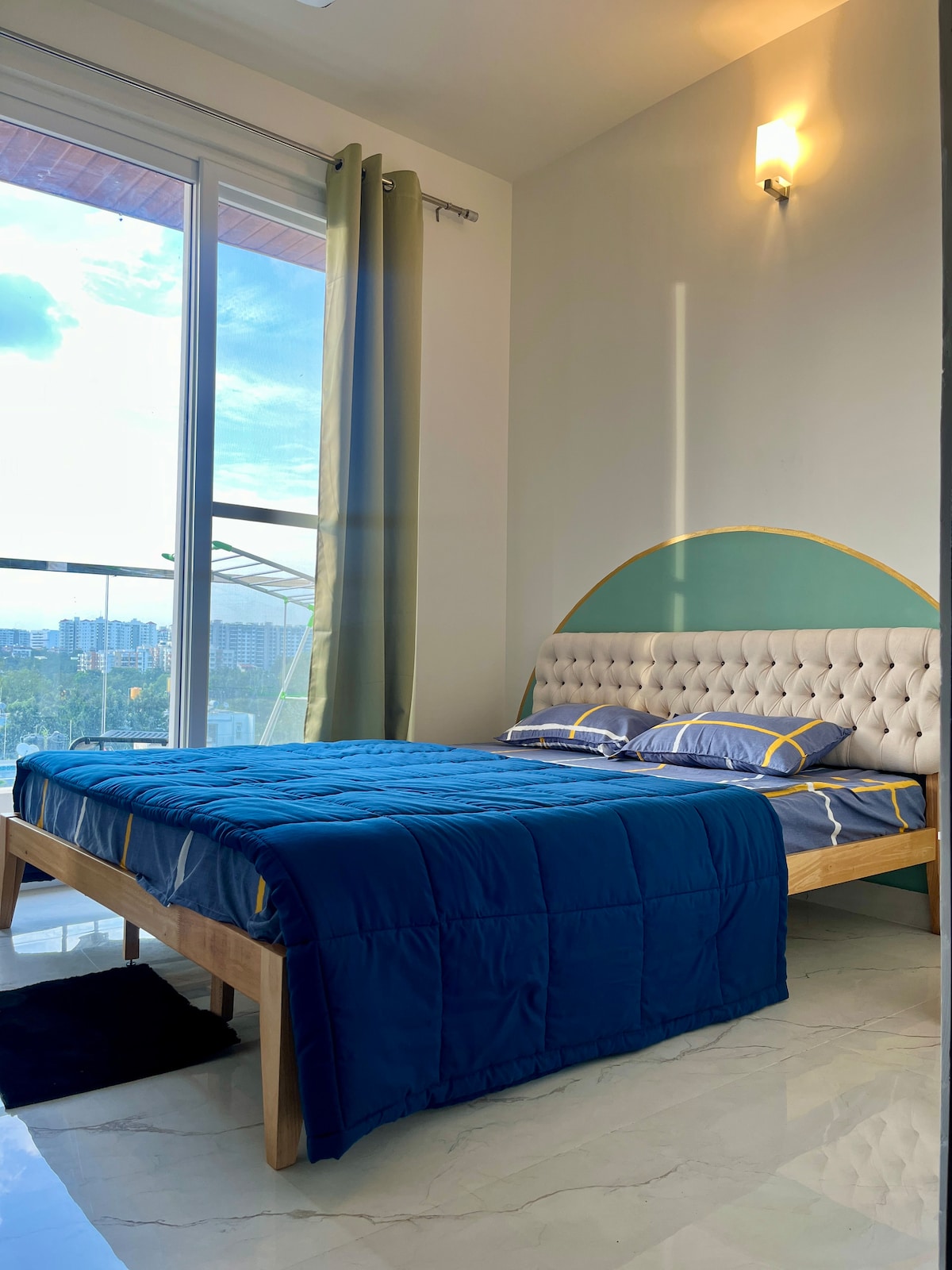 The bedroom features a spacious bed with a padded headboard and a blue quilt. Large windows allow natural light to fill the room, while curtains frame the view of the cityscape beyond. A soft rug lies beneath the bed, enhancing comfort.