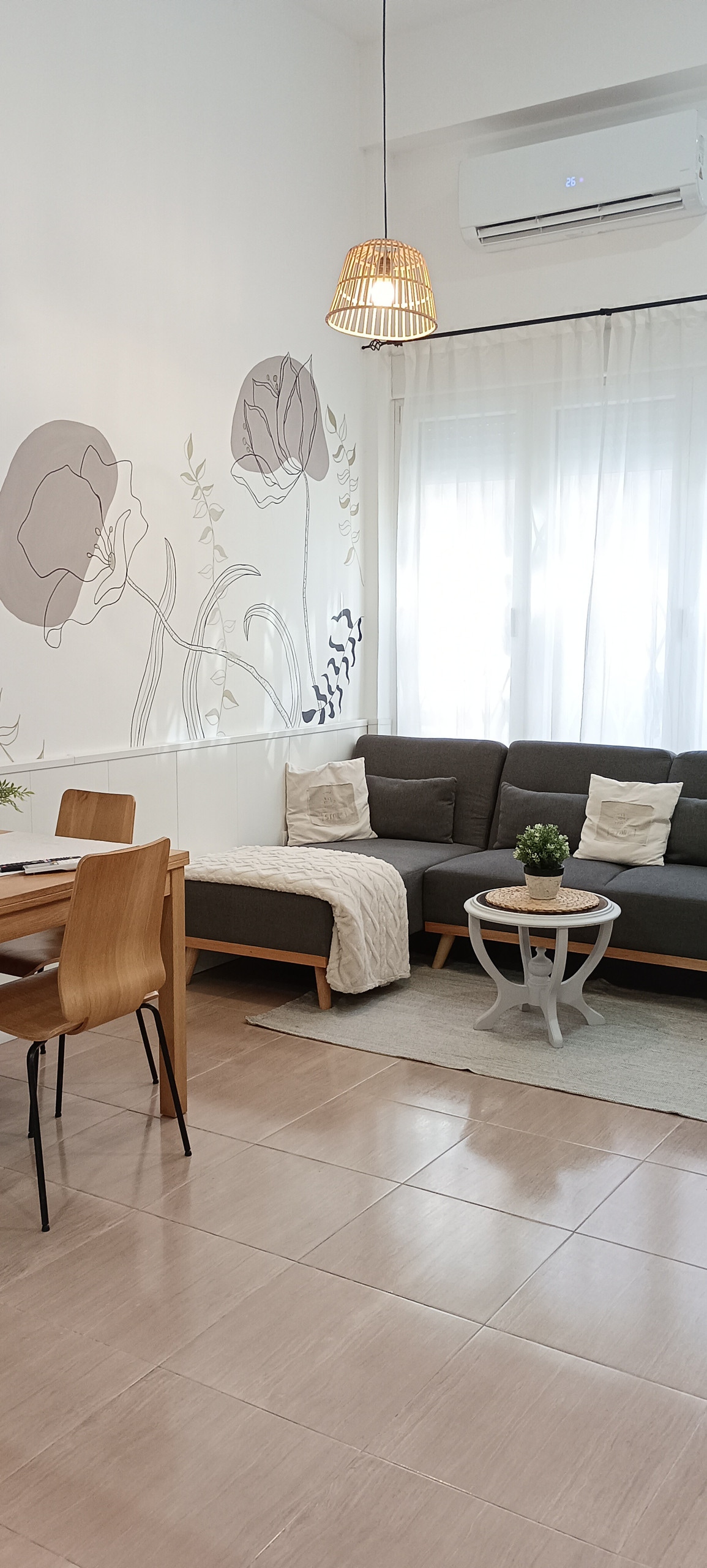 A bright living area features a gray sectional sofa, paired with decorative pillows. A small round coffee table sits in front, complemented by a plant. A dining table with wooden chairs is positioned nearby, against a backdrop of floral wall art and natural light filtering through sheer curtains.
