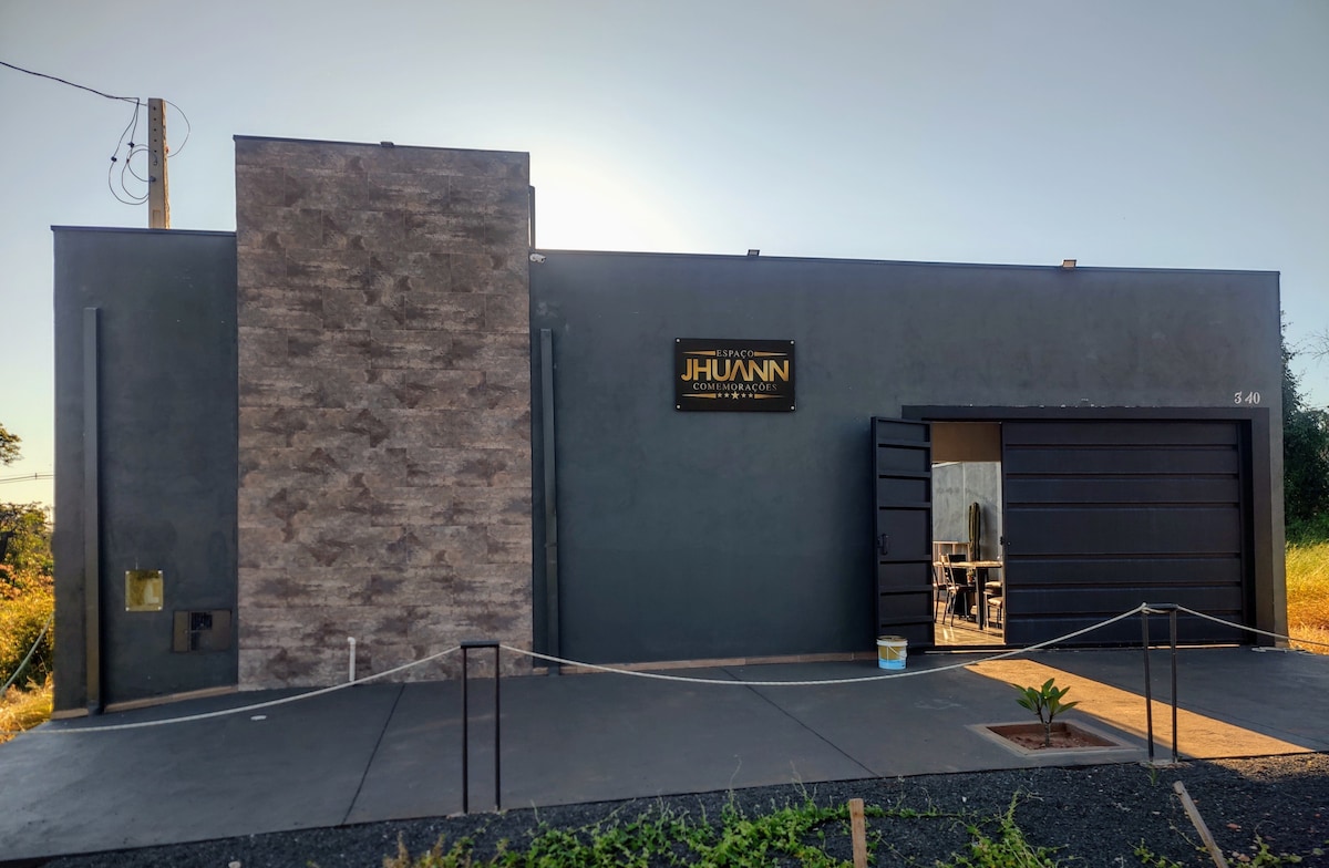 The exterior of the modern residence features a neutral color palette with a textured stone accent wall. A large garage door is visible, alongside a clean pathway leading to the entrance. Lush greenery borders the property, enhancing the welcoming atmosphere.