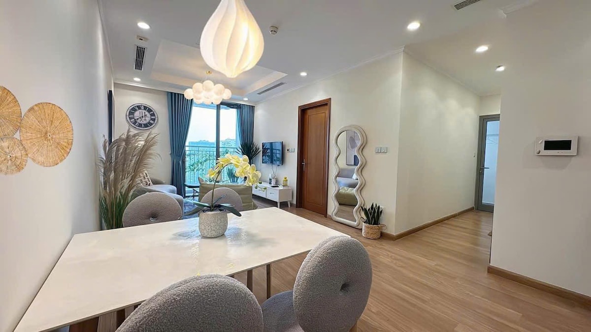 The living area is elegantly arranged, featuring a dining table with plush chairs. A soft color palette is present, complemented by a decorative lamp above. Nearby, a lounge space includes a large window with curtains and potted plants, enhancing the bright and airy atmosphere.