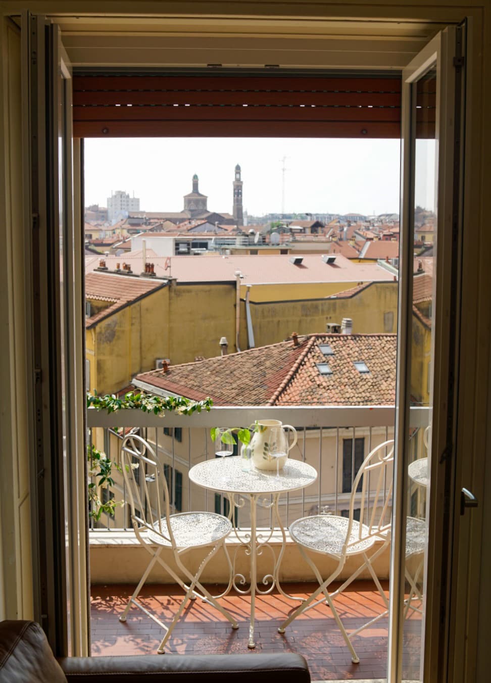 A balcony is framed by open glass doors, revealing a charming view of rooftops and distant church towers. A small round table with two delicate white chairs is positioned on the balcony, enhancing the inviting outdoor space.