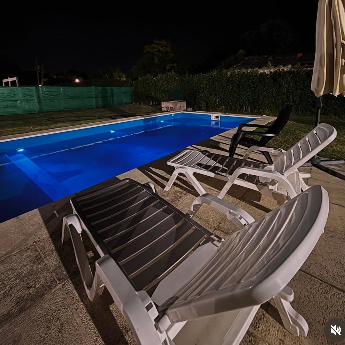 A well-lit swimming pool glows in deep blue under the night sky. Two lounge chairs are arranged beside the pool, offering space for relaxation. A neatly trimmed hedge surrounds the area, providing privacy and a sense of tranquility.