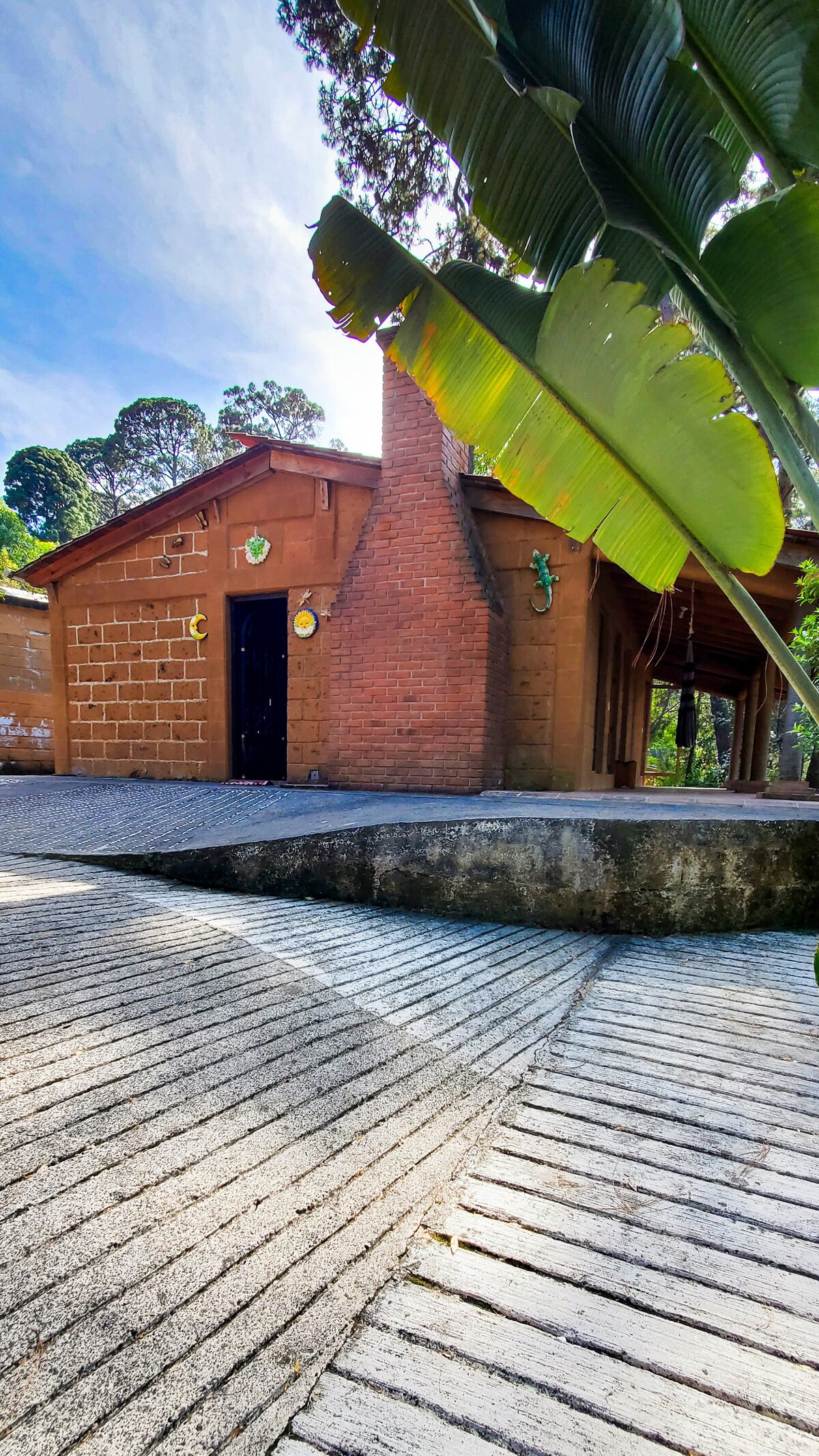 A rustic cabin is set among lush greenery, featuring earthy brick walls and a prominent chimney. A stone walkway leads to the entrance, with a covered porch extending alongside the building. Decorative elements, including wall art, add character to the exterior.