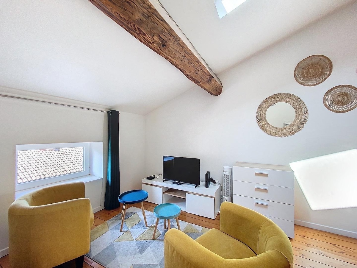 A bright living area features two yellow upholstered chairs facing a modern white TV unit with a flat-screen television. A light area rug lays on the wooden floor, complemented by three decorative mirrors and a skylight that adds natural light to the space.