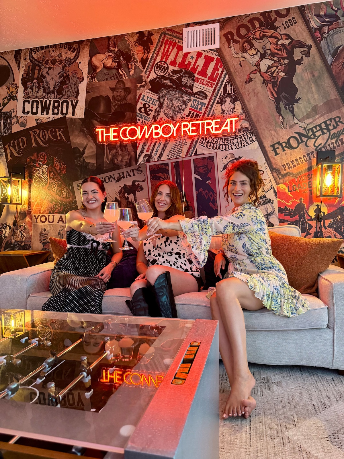 A stylish living area is showcased with Western-themed decor, featuring an inviting sofa and a decorative coffee table. Bright lights illuminate a neon sign that reads 'THE COWBOY RETREAT.' Eclectic art covers the walls, enhancing the vibrant atmosphere.