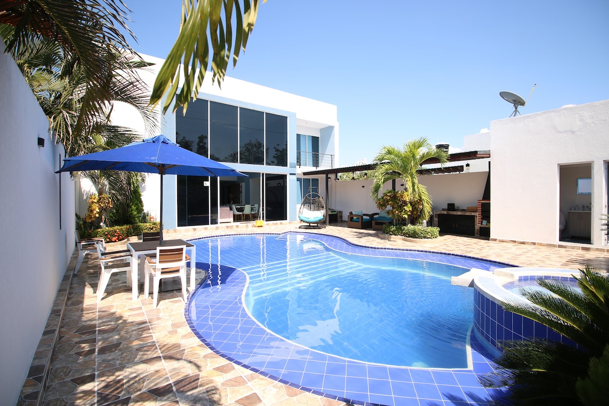 An outdoor area features a large blue swimming pool surrounded by palm trees and modern landscaping. Patio furniture, including a table and chairs, is positioned under a blue umbrella, offering shaded seating. A glass-sided building can be seen in the background, enhancing the inviting atmosphere.