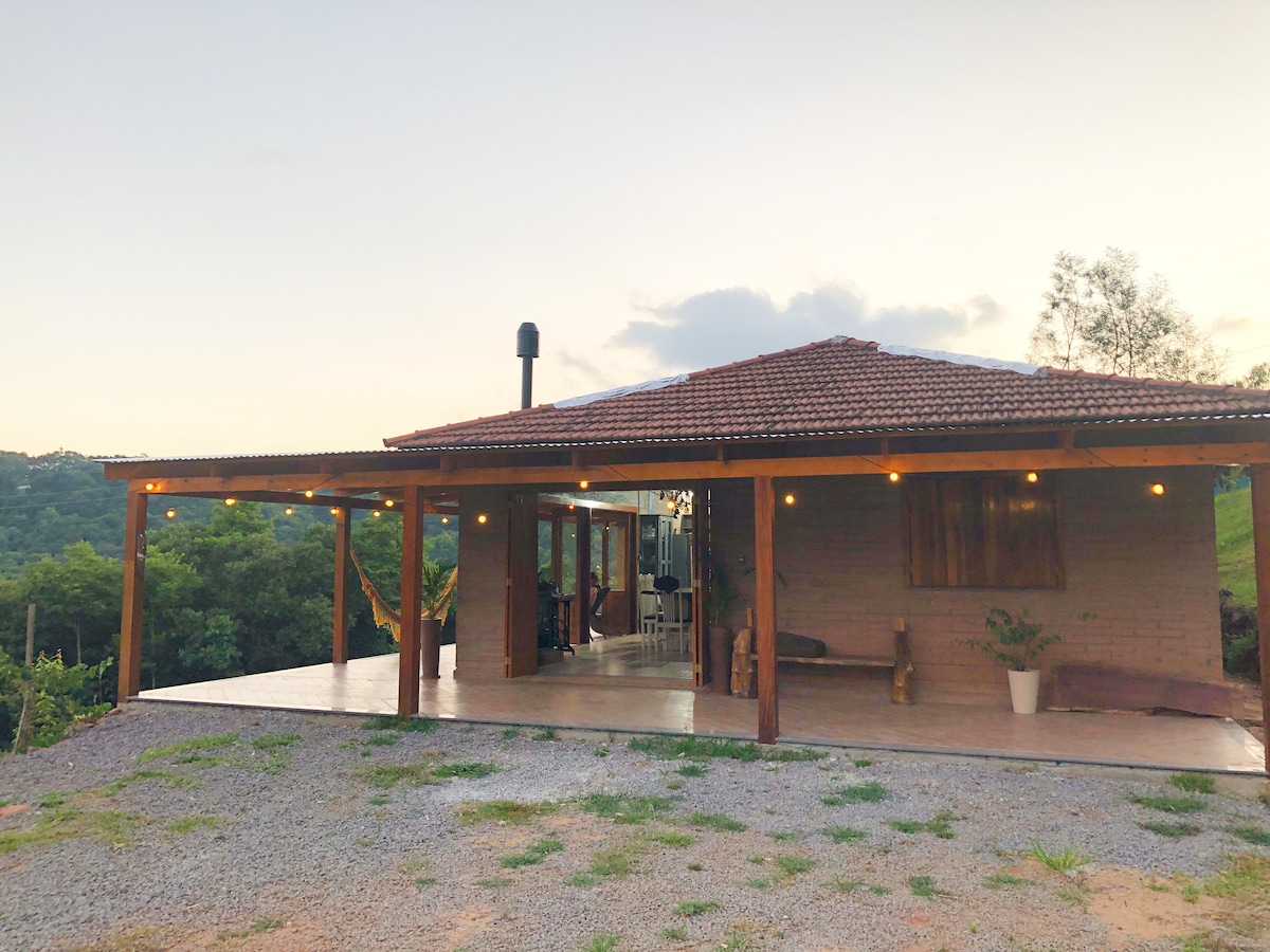A charming exterior of a rustic house features a covered porch with wooden beams and soft lighting. Surrounding greenery is visible, offering a natural backdrop. Large sliding doors reveal glimpses of the inviting interior space. A gravel path leads to the entrance.