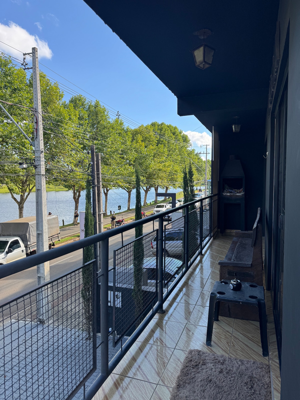 A balcony features a scenic view of the lake and tree-lined street. Seating is available with a bench and small table. The space is illuminated by natural light, enhancing the outdoor experience in a serene setting.