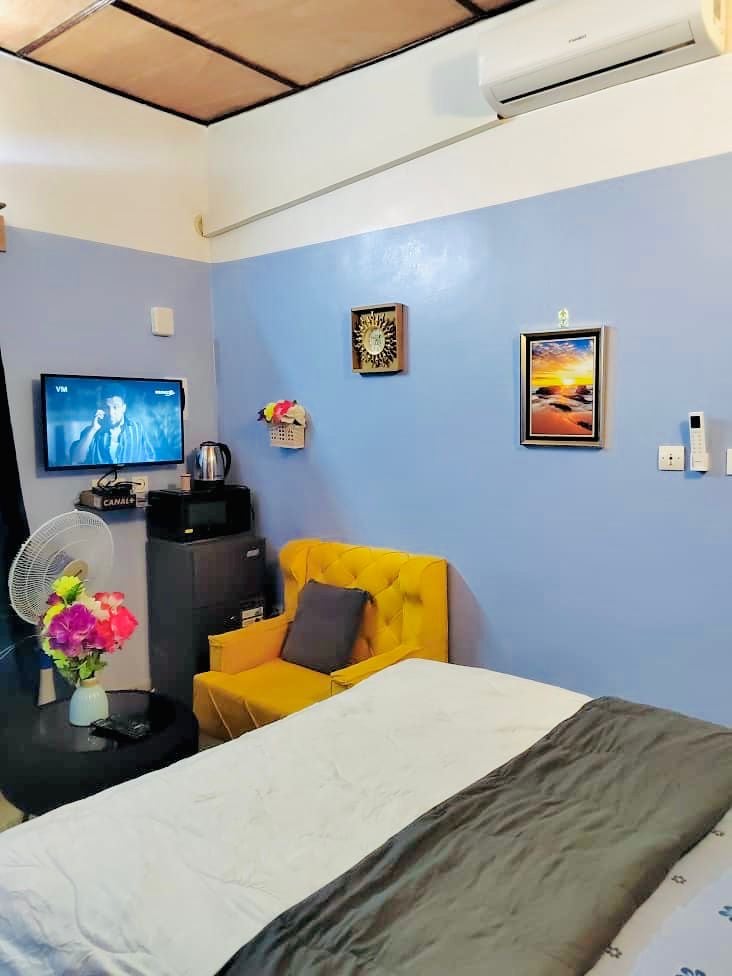 A small room features a spacious bed with a gray comforter, accented by a bright yellow armchair nearby. A wall-mounted television is visible, along with a fridge and a kettle. A fan circulates air, while framed artwork adds color to the blue walls.