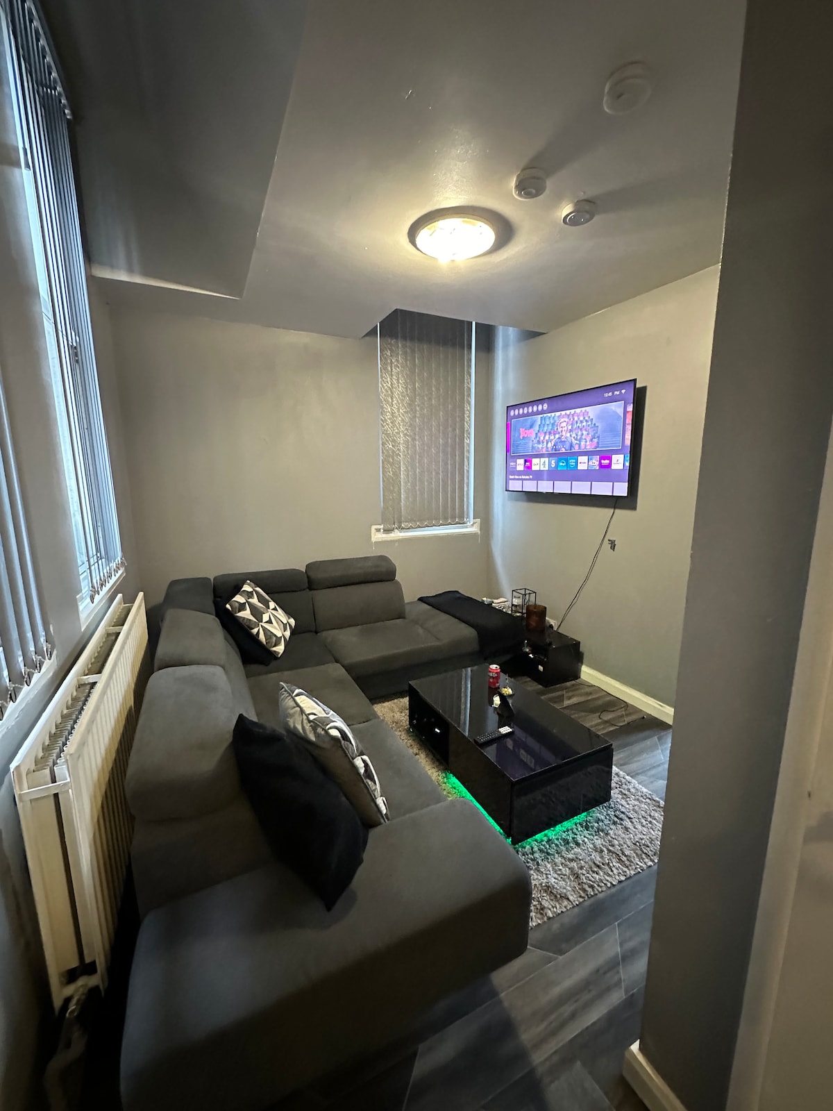 A compact living area features a gray sectional sofa adorned with black accent pillows. A sleek coffee table with subtle lighting sits in front of the sofa. A wall-mounted television displays colorful visuals, and soft lighting adds to the inviting atmosphere.
