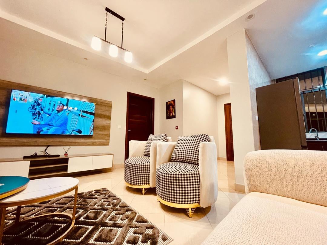 A living area is shown with two stylish armchairs featuring a checkered pattern. A spacious cream sofa faces a modern entertainment center, which houses a Hisense 4K TV. Soft lighting from a pendant lamp illuminates the neutral decor, creating a comfortable and inviting environment.