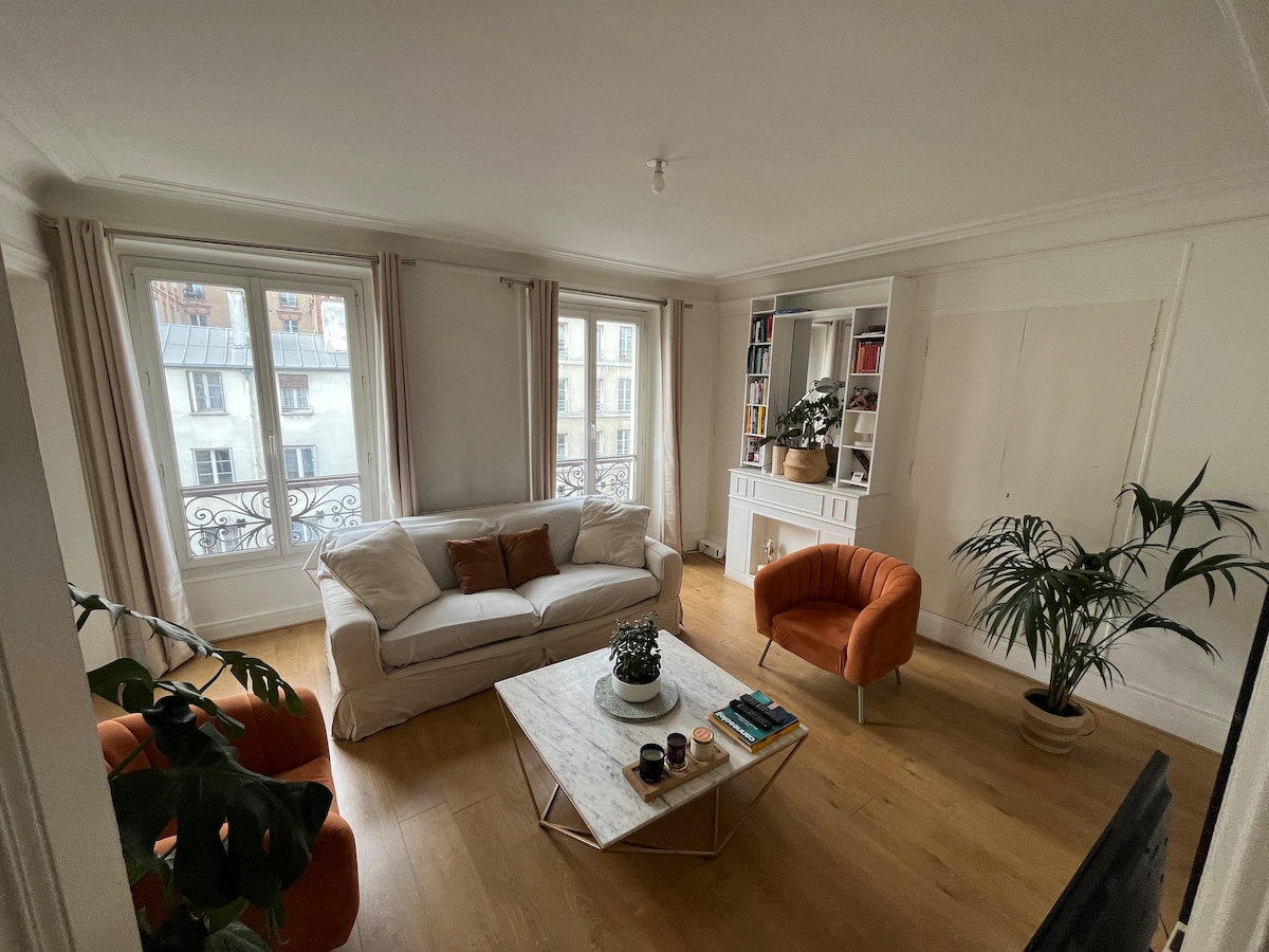 A spacious living area is filled with natural light, featuring a neutral-coloured sofa and two orange armchairs. A marble coffee table sits at the center, complemented by a small plant and books on top. Large windows with light curtains provide views of the outside.