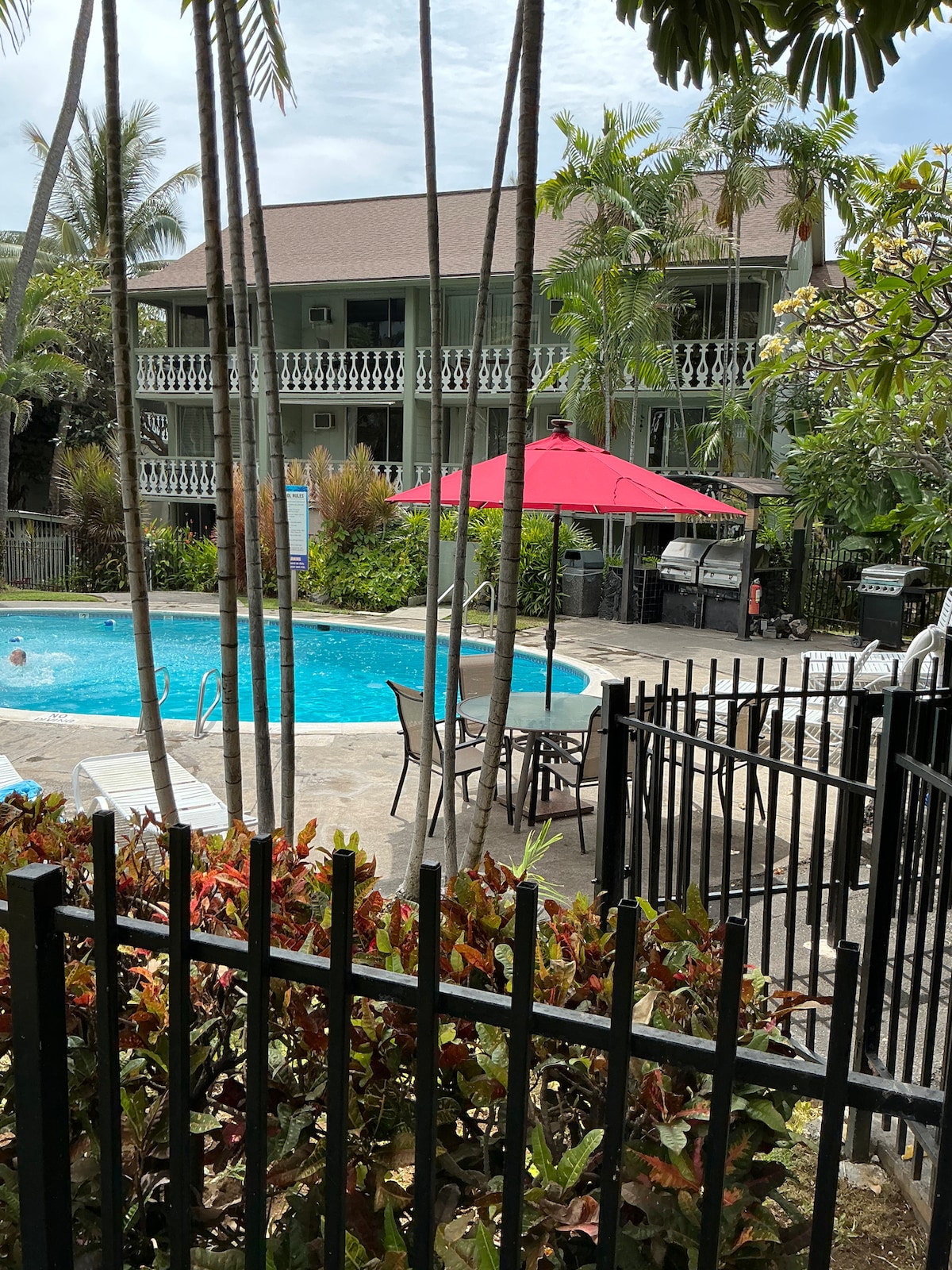 A tranquil pool area is surrounded by lush greenery and palm trees. A red umbrella provides shade over a small seating arrangement near the water. The two-story building in the background features decorative railings and is set within a serene outdoor environment.