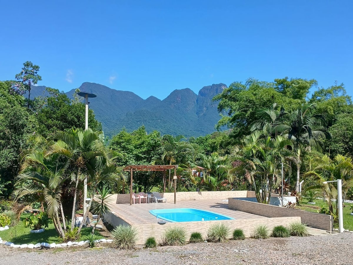 A serene outdoor area is captured, featuring a clear blue pool surrounded by lush greenery and tropical plants. Wooden structures provide shade and seating, with mountains rising in the background against a bright sky.