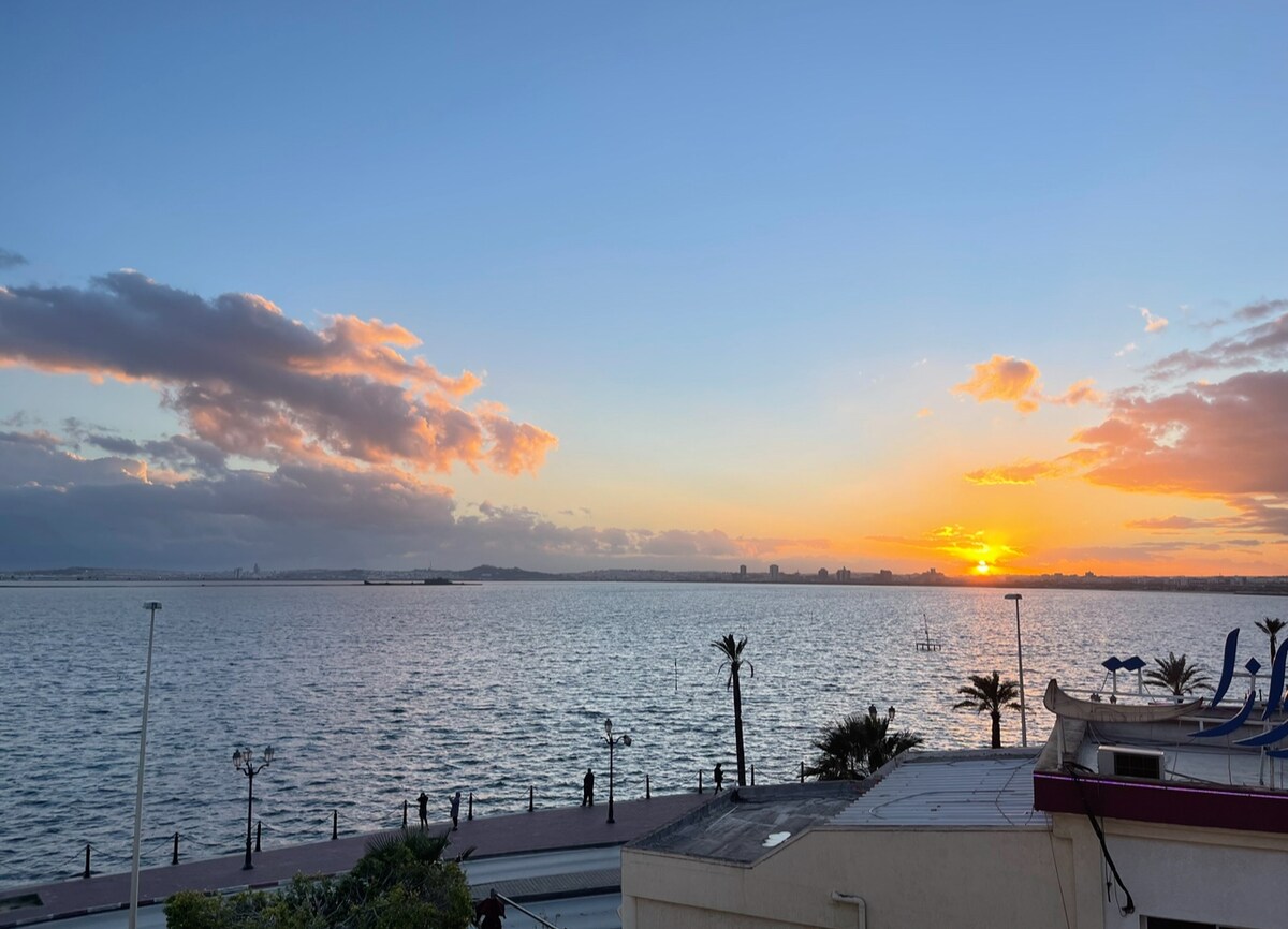 A scenic view of the sun setting over the water, casting warm hues across the sky. Silhouetted palm trees and a pathway can be seen, with people enjoying the waterfront. The gentle waves reflect orange and pink tones, creating a serene atmosphere.