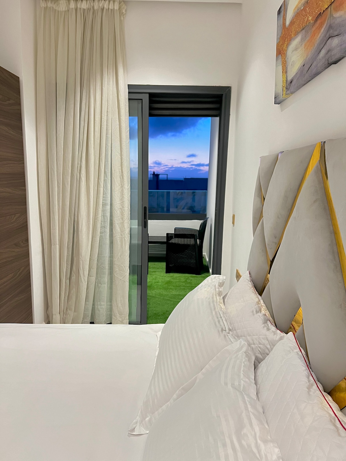 A spacious bedroom is depicted with a plush bed adorned with multiple pillows. Sheer curtains frame the doorway leading to a private balcony, where a glimpse of outdoor furniture and a sky filled with soft hues can be seen.