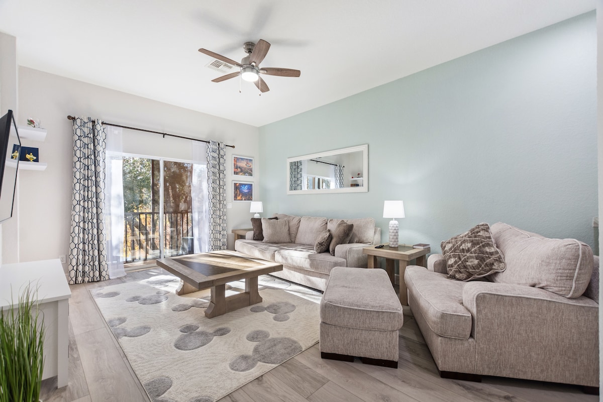 The living area features a spacious sofa and a cozy armchair arranged around a wooden coffee table. Light is provided by two table lamps on side tables. A large mirror and sliding glass doors open to a balcony, complementing the soft-coloured walls and patterned area rug.