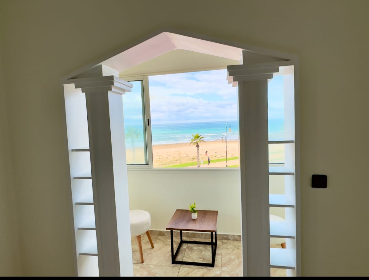 A bright living space is framed by arched architectural elements, with a view showcasing the beach and ocean. A small table is positioned near a chair, and natural light fills the room through a large window, enhancing the open atmosphere.