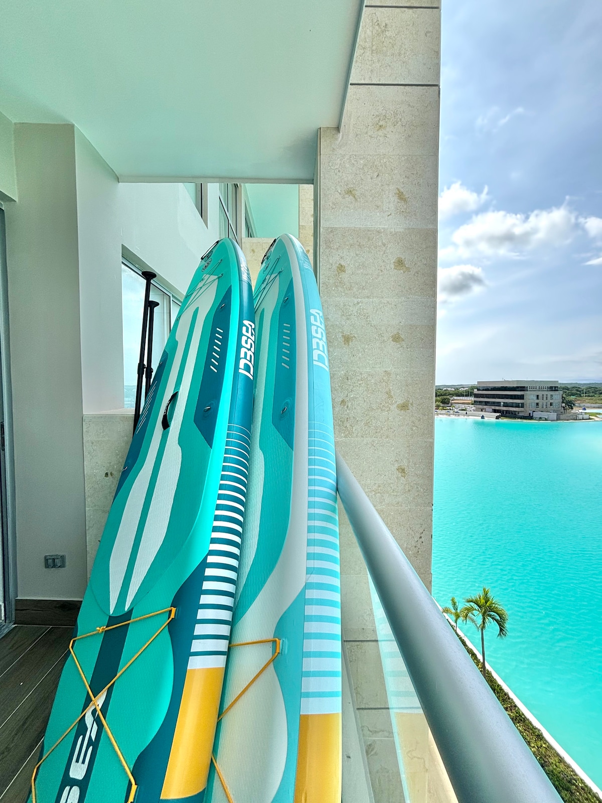 Two paddleboards are secured on a balcony railing, showcasing their vibrant colors and designs. The clear turquoise water of the lagoon is visible in the background, complemented by a clear sky and palm trees, creating a serene aquatic setting.