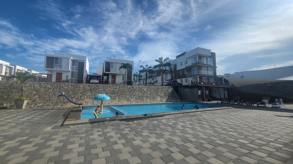 A spacious pool area is surrounded by a smooth stone wall, featuring a large swimming pool with a vibrant blue color. Comfortable lounge chairs are arranged along the poolside, and modern buildings are visible in the background, complemented by palm trees under a clear sky.