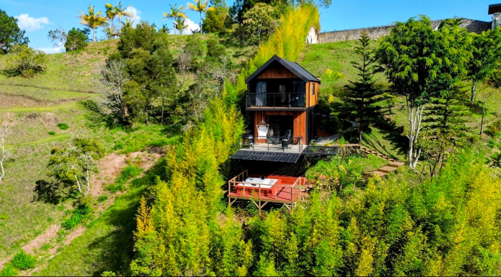 The cabin is nestled among lush greenery, surrounded by tall bamboo and trees. A spacious deck with outdoor seating is visible, providing an inviting space to enjoy the natural surroundings. The two-story structure features a sleek design with a balcony overlooking the scenic landscape.