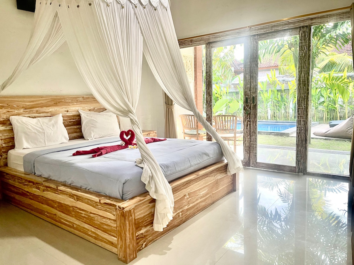 A spacious bed is framed by a wooden structure, adorned with white drapery. Large glass doors offer a view of the lush outdoor environment, which includes greenery and a swimming pool. Sunlight illuminates the room, reflecting off polished tile flooring.