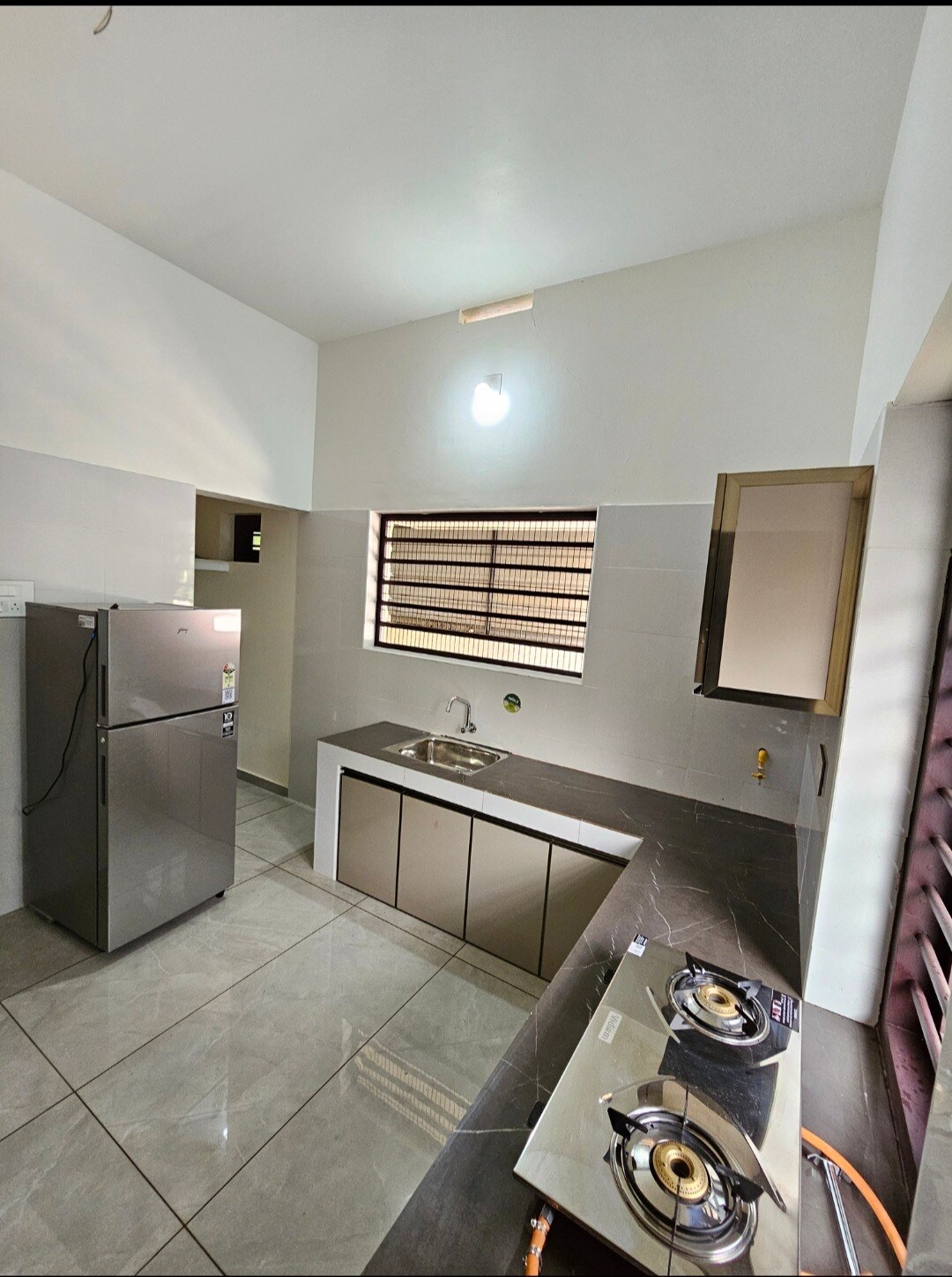 The kitchen features modern cabinetry with a dark countertop and a stainless steel sink. A gas stove is positioned prominently, and a refrigerator stands nearby. Natural light enters through a window with horizontal grills, illuminating the space.