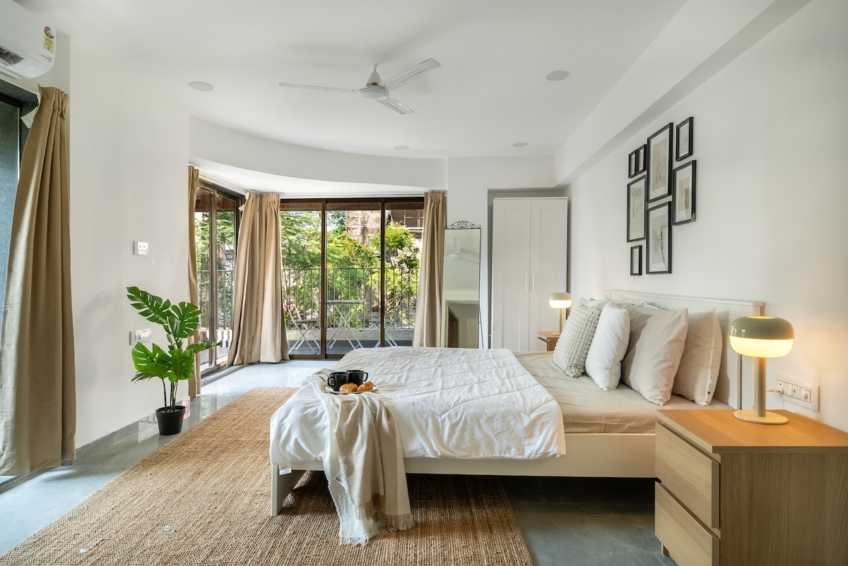 A spacious bedroom features large glass doors that lead to a private balcony. A neutral color palette is complemented by tasteful decor, including framed artwork and a leafy plant. The cozy bed is accessorized with soft linens, and a modest seating area offers additional comfort.