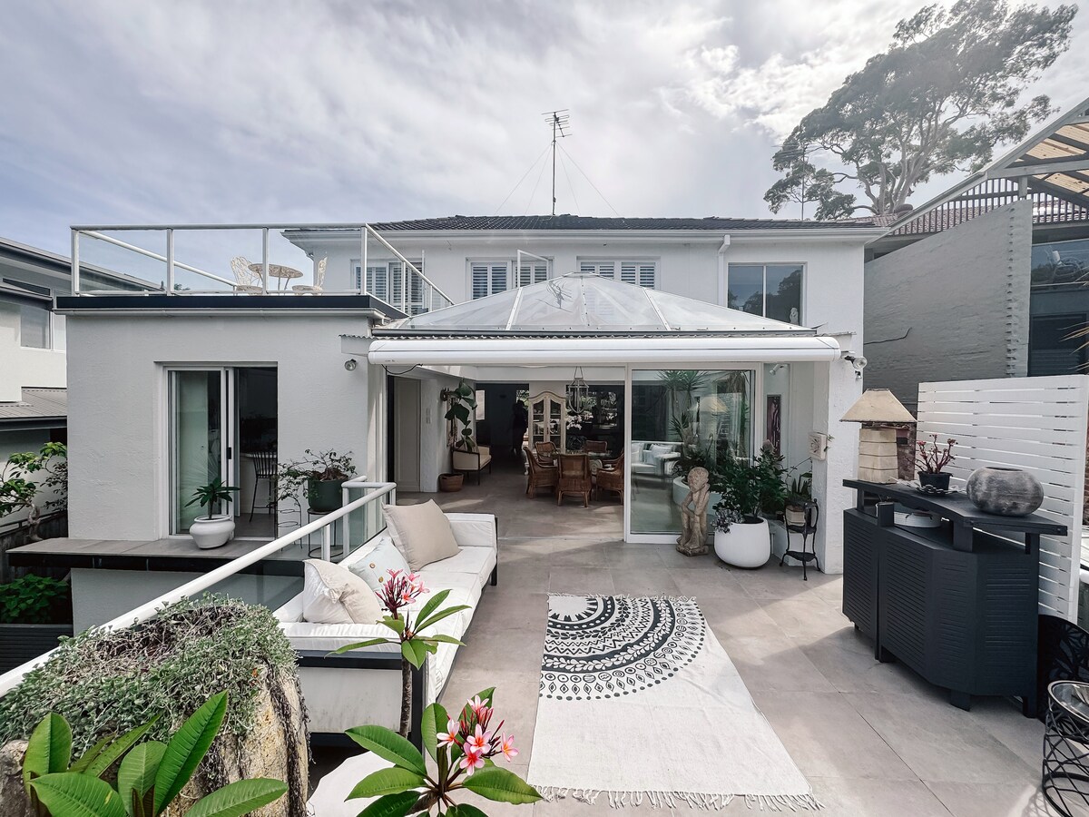 The outdoor area showcases a spacious patio with a comfortable seating arrangement, featuring neutral-toned cushions. A large glass-roofed space connects to the interior, offering natural light. Lush plants and decorative elements enhance the inviting atmosphere, while a patterned rug adds texture to the outdoor setting.