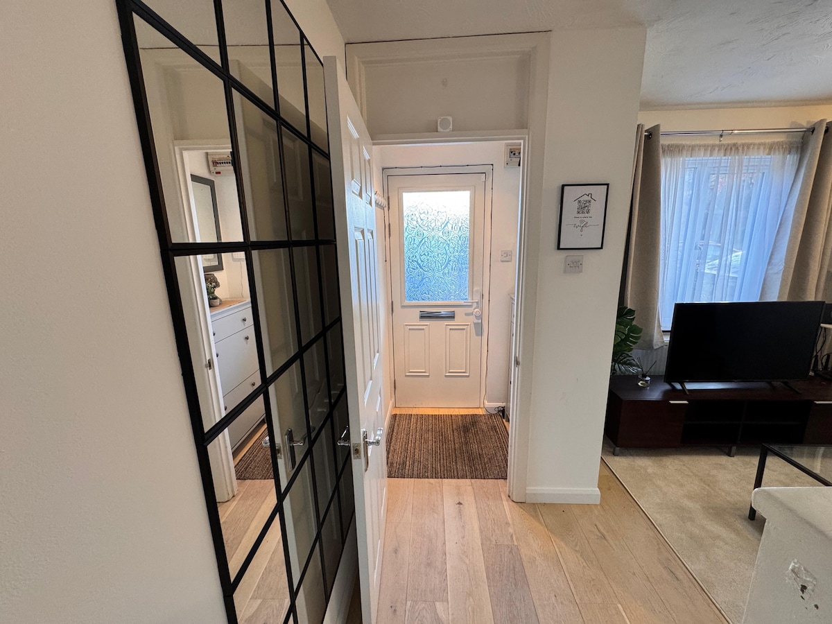 An entryway is visible, featuring a glass-paned door with a textured surface, allowing light to enter. A mirror is mounted on the wall, reflecting the hallway. Natural wood flooring extends towards a spacious living area, where a flat-screen TV is positioned near a large window dressed with sheer curtains.
