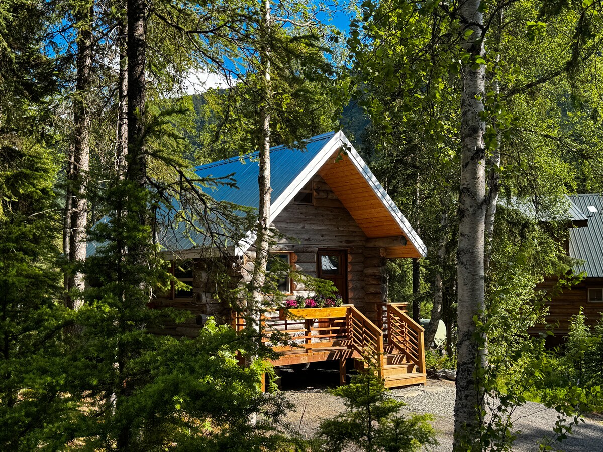 A cozy log cabin is surrounded by tall trees and lush greenery. The cabin features a peaked roof and a welcoming front porch with wooden railings. Sunlight filters through the branches, highlighting the natural setting and vibrant flowers by the windows.