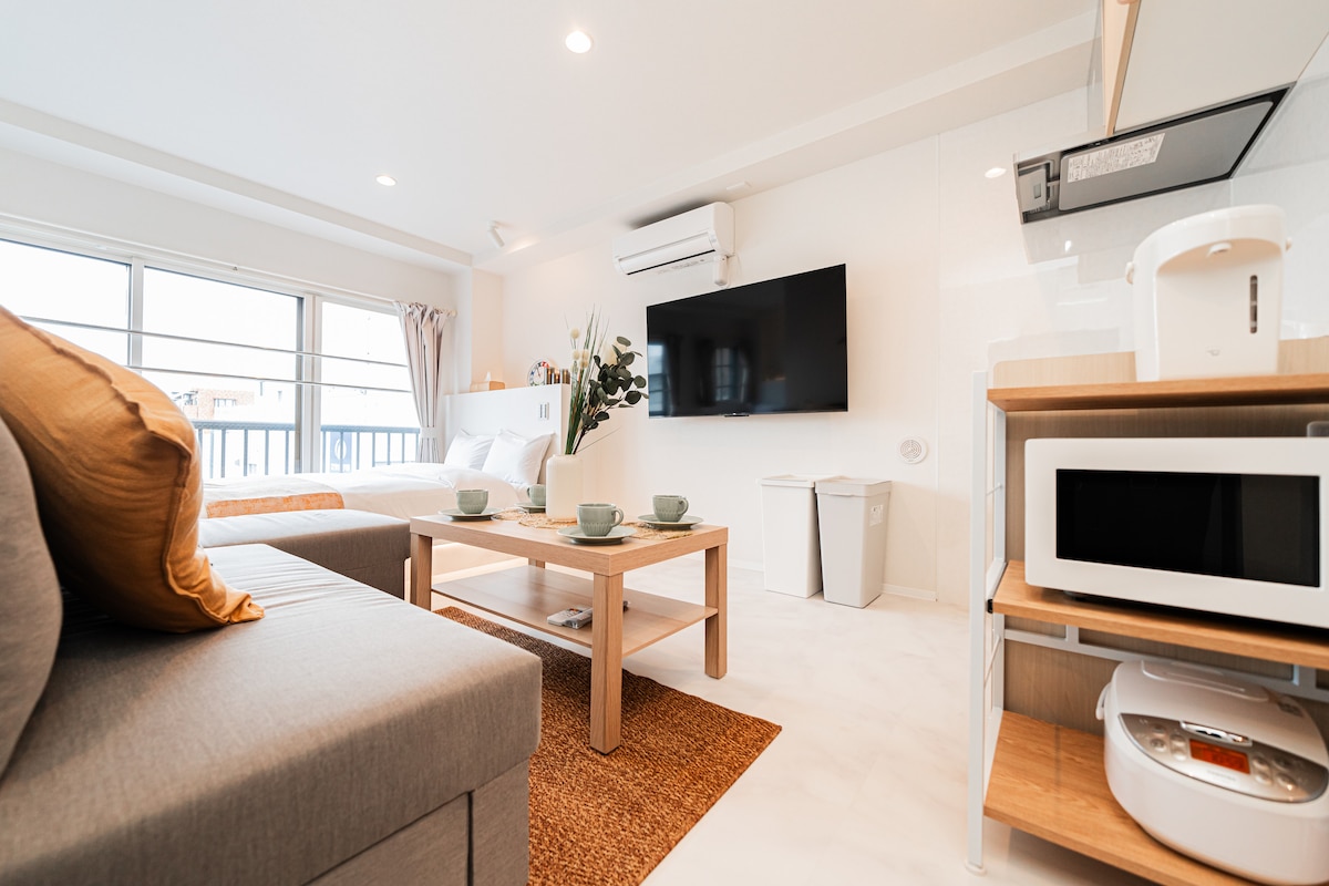 The living area is highlighted by a cozy sofa and a modern coffee table. A flat-screen television is mounted on the wall. Natural light fills the space through large windows, while a kitchenette is equipped with a microwave and essential dining items. Decorative plants add a touch of freshness.