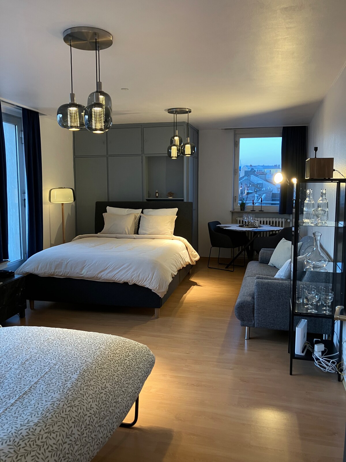 A cozy studio space features a spacious bed with soft bedding, accompanied by a compact dining table and two chairs. A comfortable seating area is present, alongside a glass display shelf showcasing decorative items. Large windows allow natural light to brighten the room.