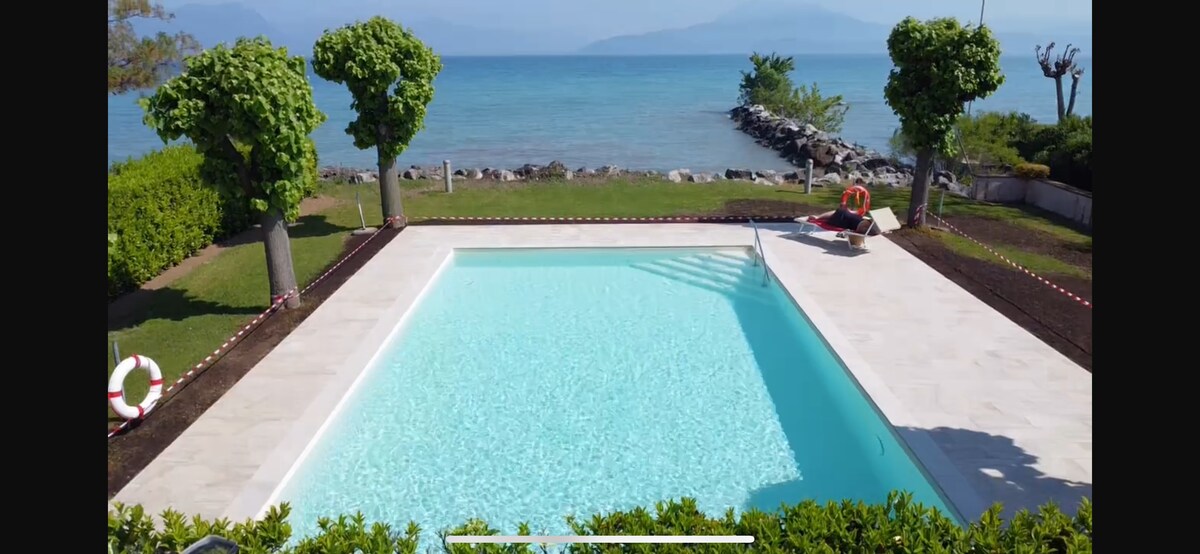 A serene swimming pool features clear turquoise water, surrounded by manicured green grass and stone pathways. Lush trees provide shade, and a stone jetty is visible along the coastline. The backdrop showcases tranquil lake views and distant mountains.