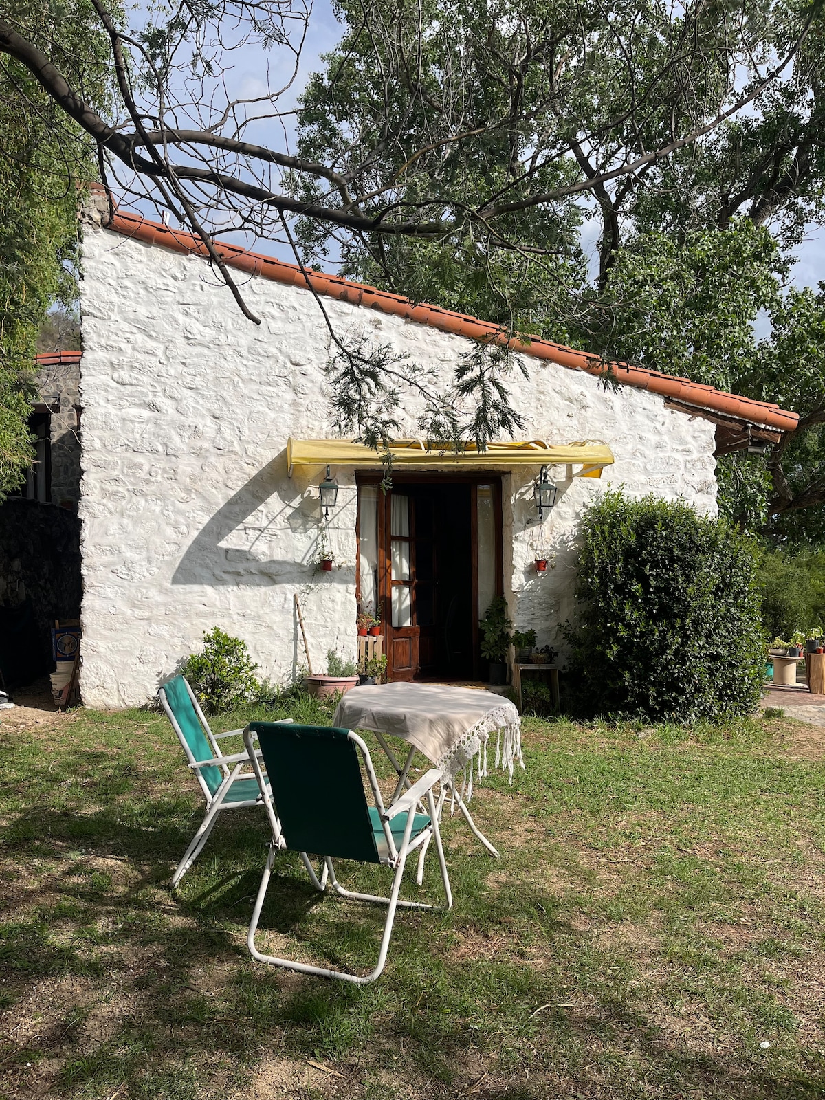 A charming exterior view of the refuge showcases a stone wall structure with a terracotta roof. A yellow awning provides shade above the entrance. A small table is set with two folding chairs on the grassy area, surrounded by lush greenery.