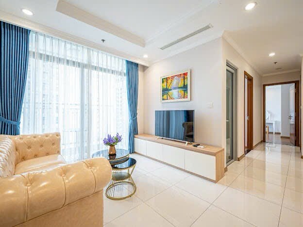 A stylish living area features a beige tufted sofa and a sleek black coffee table. Floor-to-ceiling windows are adorned with elegant blue curtains, allowing natural light to fill the space. A flat-screen TV is mounted on a minimalistic white cabinet, enhancing the modern design.