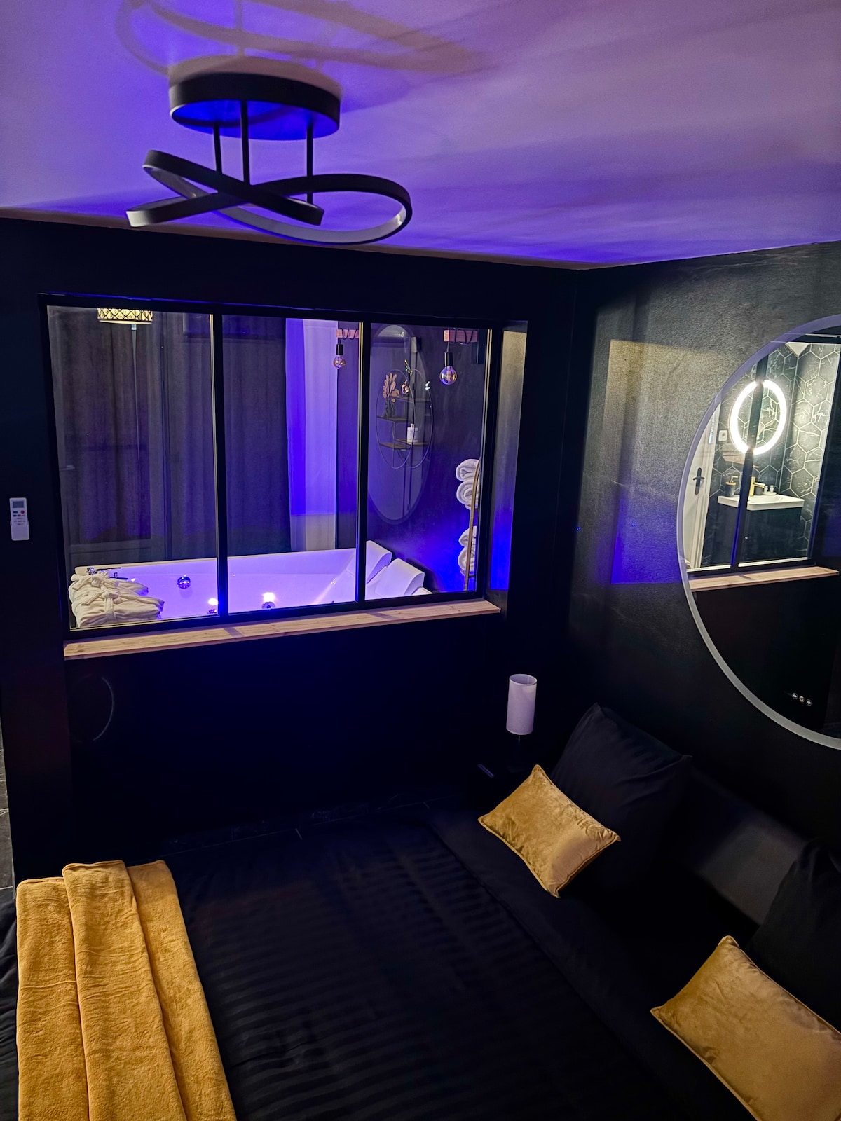 The intimate sleeping area features dark walls and a comfortable sleeping surface, complemented by soft gold accents. A large circular mirror adorns one wall, reflecting the nearby illuminated spa area visible through a glass partition, enhancing the ambiance with subtle blue lighting.